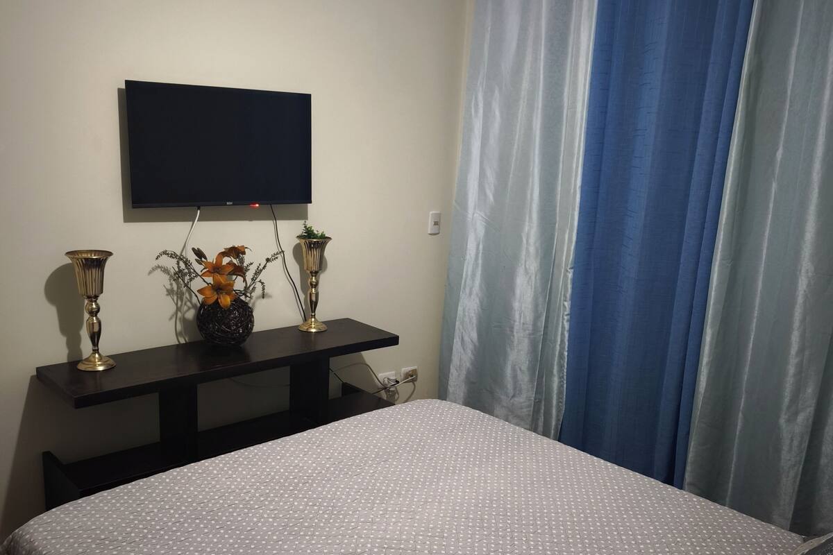 Een slaapkamer met een bed, een tv aan de muur, een klein tafeltje met decoratieve spullen en een raam met gordijnen.