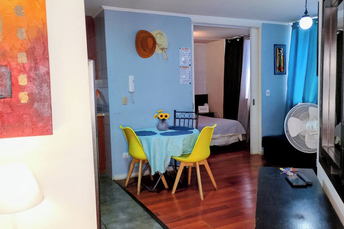 Een ruimtelijk ingerichte leefruimte met een eethoek bestaande uit een tafel en stoelen, een bed, een ventilator en een aan de muur bevestigde telefoon.