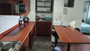 Een kleine keuken met houten kastjes, een magnetron, een spoelbak en een eethoek met een stoel.