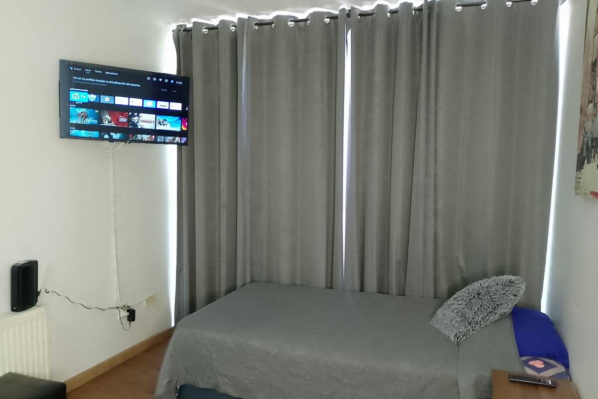 Een slaapkamer met een bed, een aan de muur bevestigde televisie en een raam met gordijnen.