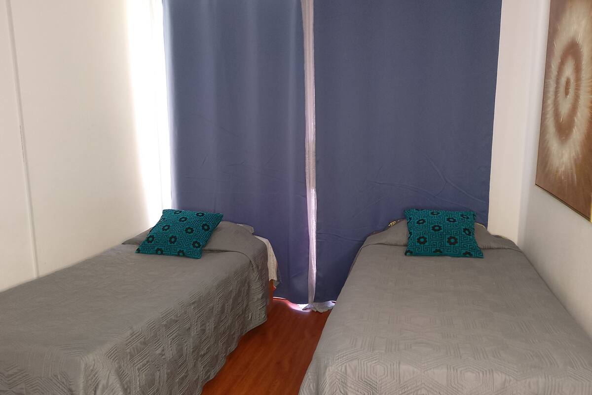 Twee eenpersoonsbedden met beddengoed met dessin, een houten vloer en een raam met gordijnen.
