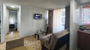 Een woonkamer met een bank, een salontafel, een aan de muur bevestigde televisie en een keuken met een spoelbak en kasten.