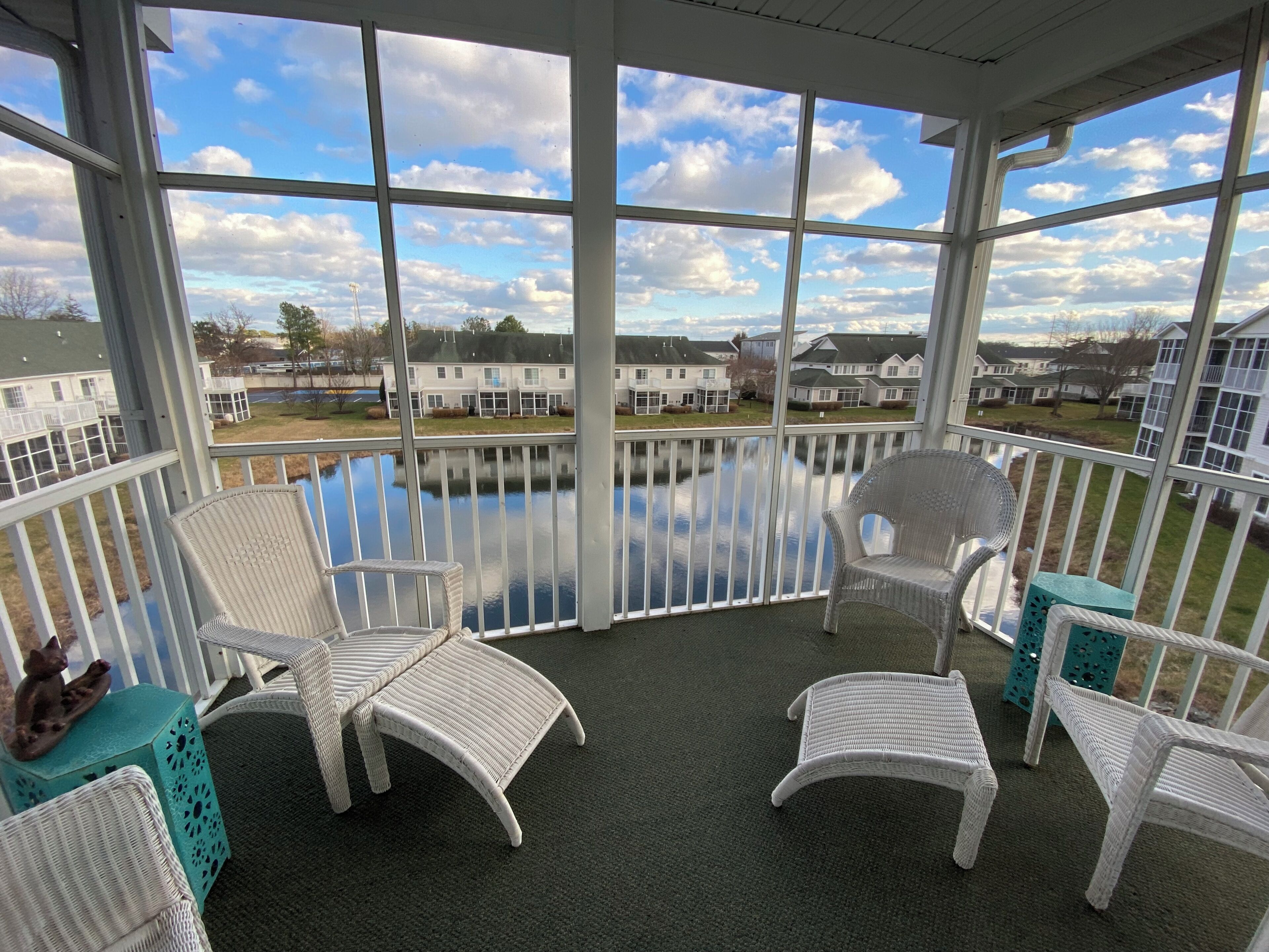 A covered balcony with white wicker chairs and a view of a residential area and a body of water.