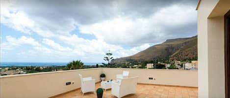 A terrace with white chairs and a table, overlooking a coastal landscape with mountains and a view of the sea.