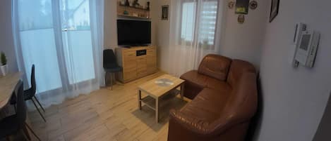 A living room with a brown leather sofa, a wooden coffee table, a TV on a wooden cabinet, and a dining area with a table and chairs.