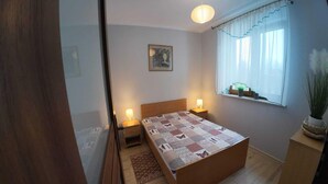 Une chambre avec un lit en bois, deux lampes de chevet, une fenêtre avec des rideaux légers, une petite plante sur le rebord de la fenêtre et un tableau encadré au mur.