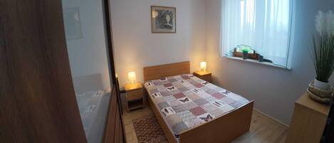 A bedroom with a wooden bed, two bedside lamps, a window with sheer curtains, a small plant on the windowsill, and a framed picture on the wall.