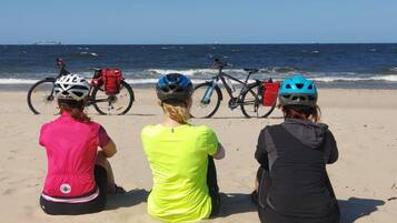 Tres personas sentadas en una playa arenosa con bicicletas y cascos, mirando al océano.