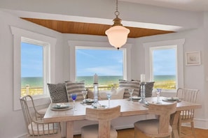 Un espace repas offrant une vue sur l’océan, avec une table en bois dressée pour quatre personnes, des murs blancs et un plafond avec des poutres apparentes.