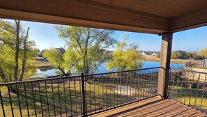A covered outdoor area with a wooden deck, metal railing, and a view of a lake and trees. - On The Pond (Granbury)