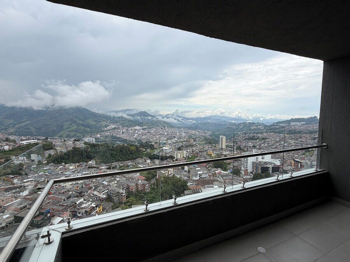 Un balcon doté d’une balustrade en métal, d’où l’on peut admirer un paysage urbain avec des bâtiments et des montagnes à l’horizon.