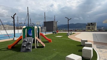 Un parque infantil con toboganes y estructuras para escalar, rodeado de césped artificial y bancos de hormigón.