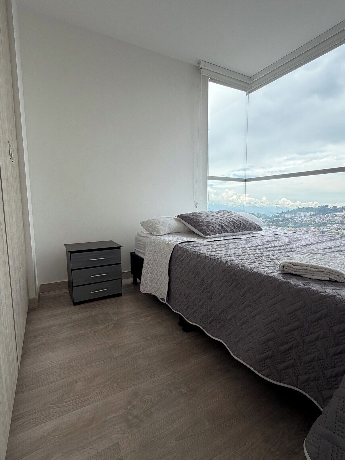 Una camera da letto con un letto, un comodino, una cassettiera e un'ampia finestra con vista sulla città.