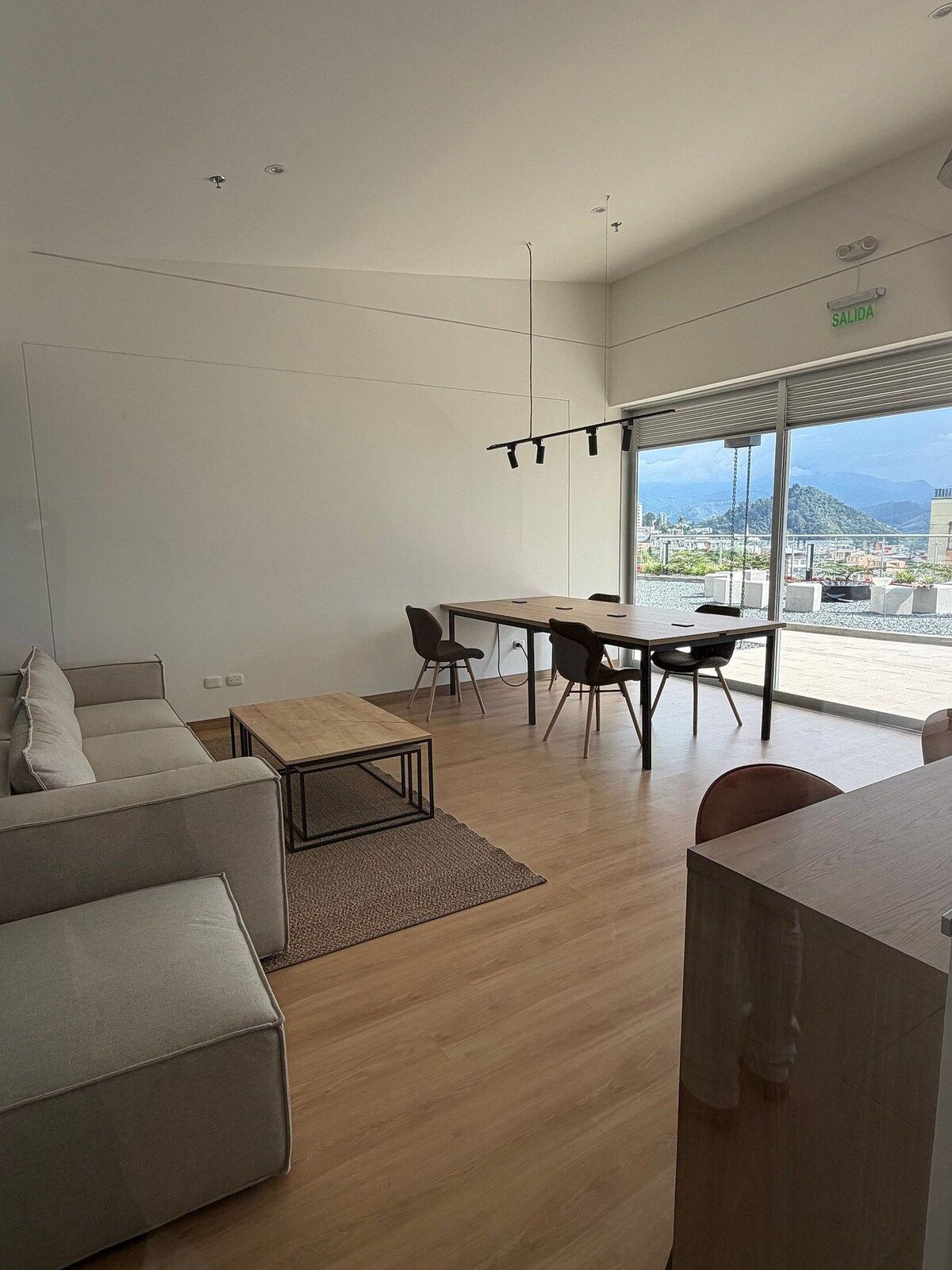 Una sala de estar moderna con un sofá, una mesa de centro y un comedor con sillas. Hay un ventanal que ofrece vistas de la ciudad.
