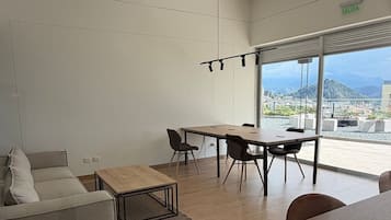 Una sala de estar moderna con un sofá, una mesa de centro y un comedor con sillas. Hay un ventanal que ofrece vistas de la ciudad.