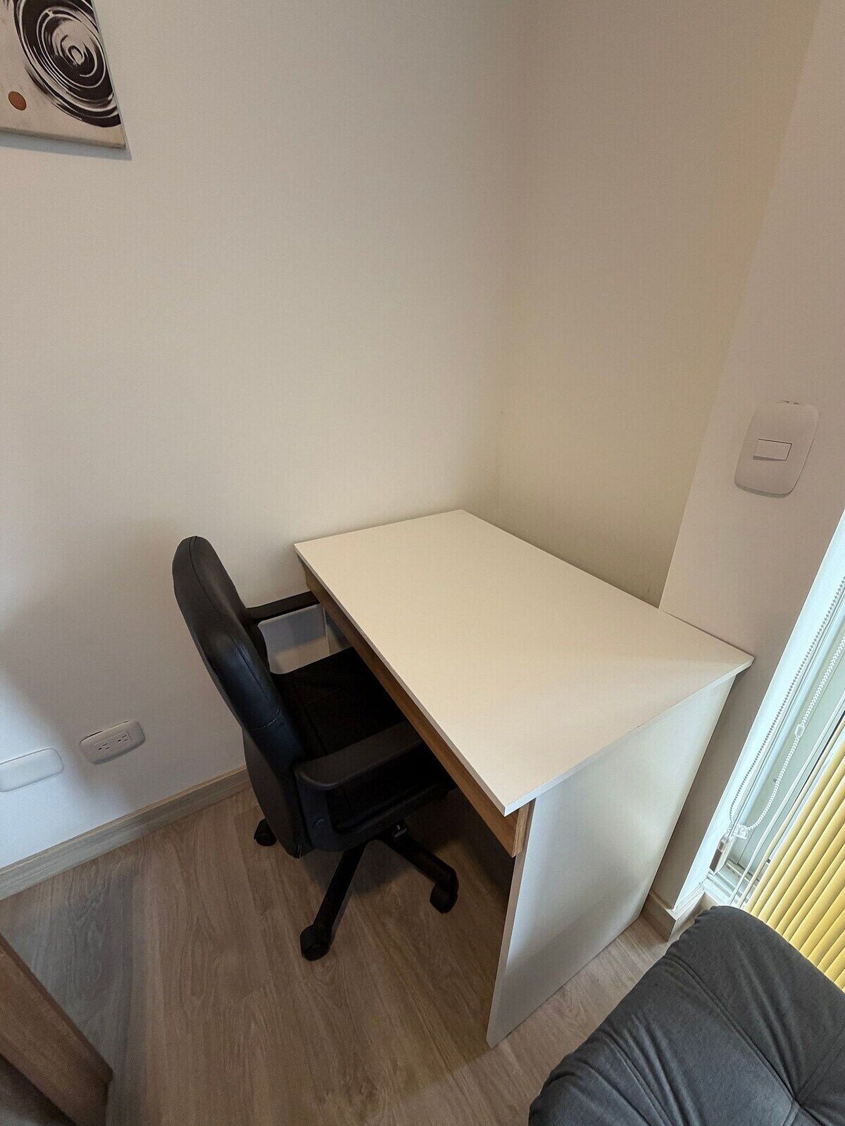 Ein kleiner, ordentlicher Arbeitsplatz mit Schreibtisch, Stuhl und einem Fenster mit Jalousien.