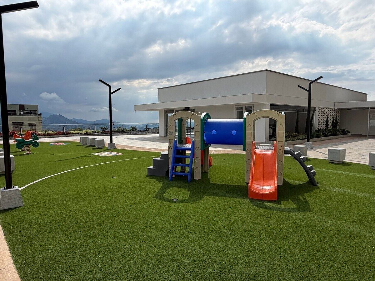 Zona de juegos al aire libre con un tobogán y estructuras para escalar, rodeada de césped artificial y un edificio al fondo.