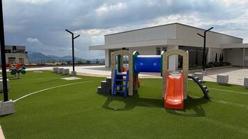 Zona de juegos al aire libre con un tobogán y estructuras para escalar, rodeada de césped artificial y un edificio al fondo.