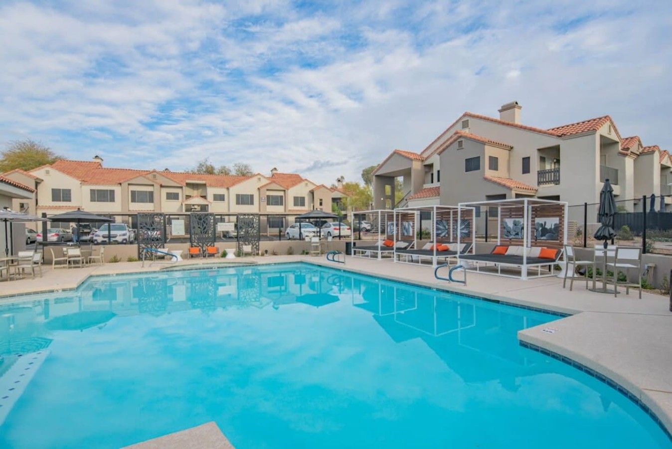 A swimming pool with lounge chairs and umbrellas, surrounded by multi-story buildings with red-tiled roofs.