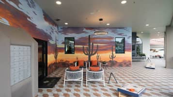 A room with a desert mural, two white wicker chairs with orange cushions, a small table, and a pool table.