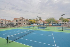 An outdoor tennis court with multiple courts, surrounded by residential buildings and a fence.