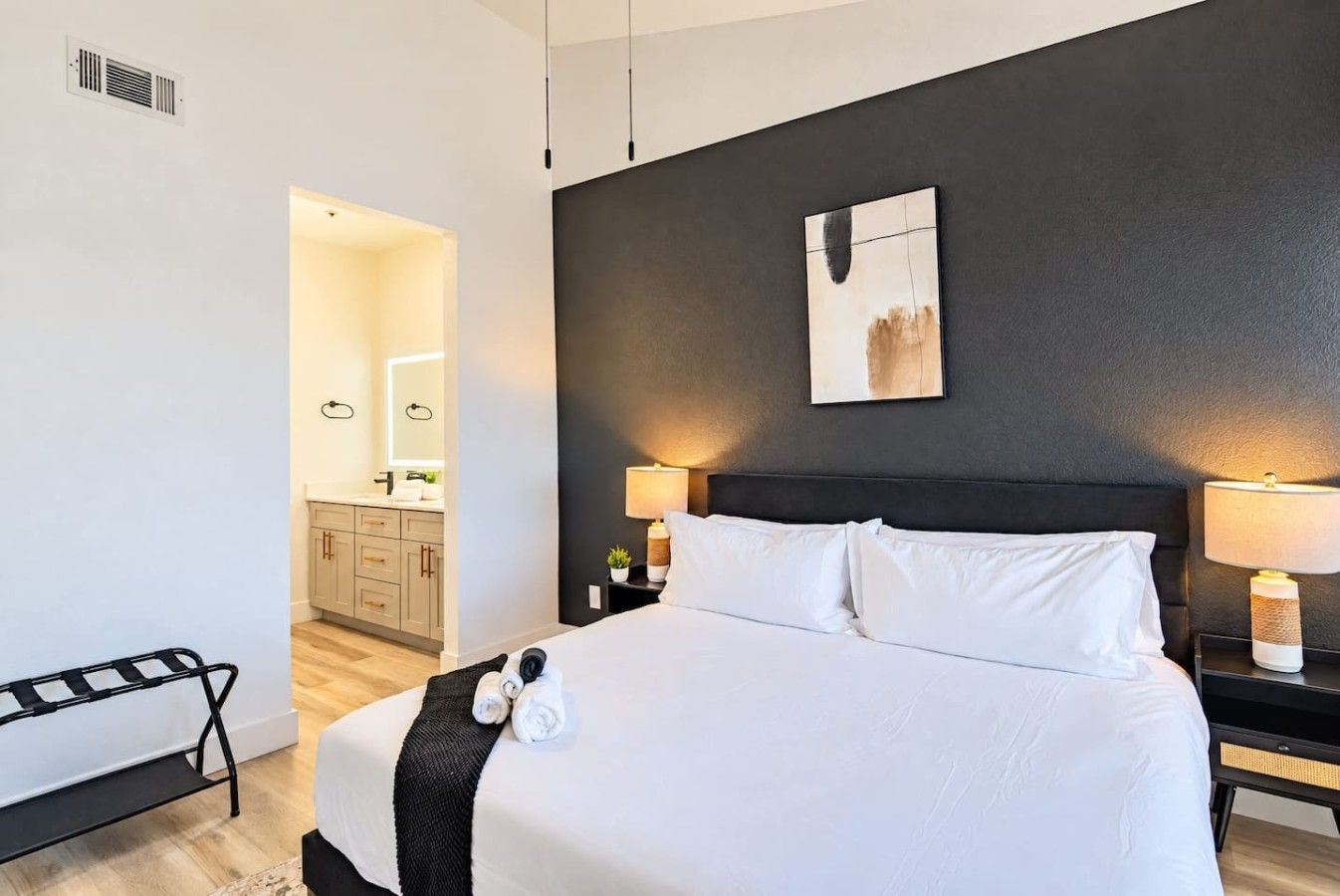 A hotel room with a large bed, bedside lamps, a headboard, a painting, and a view into the bathroom.