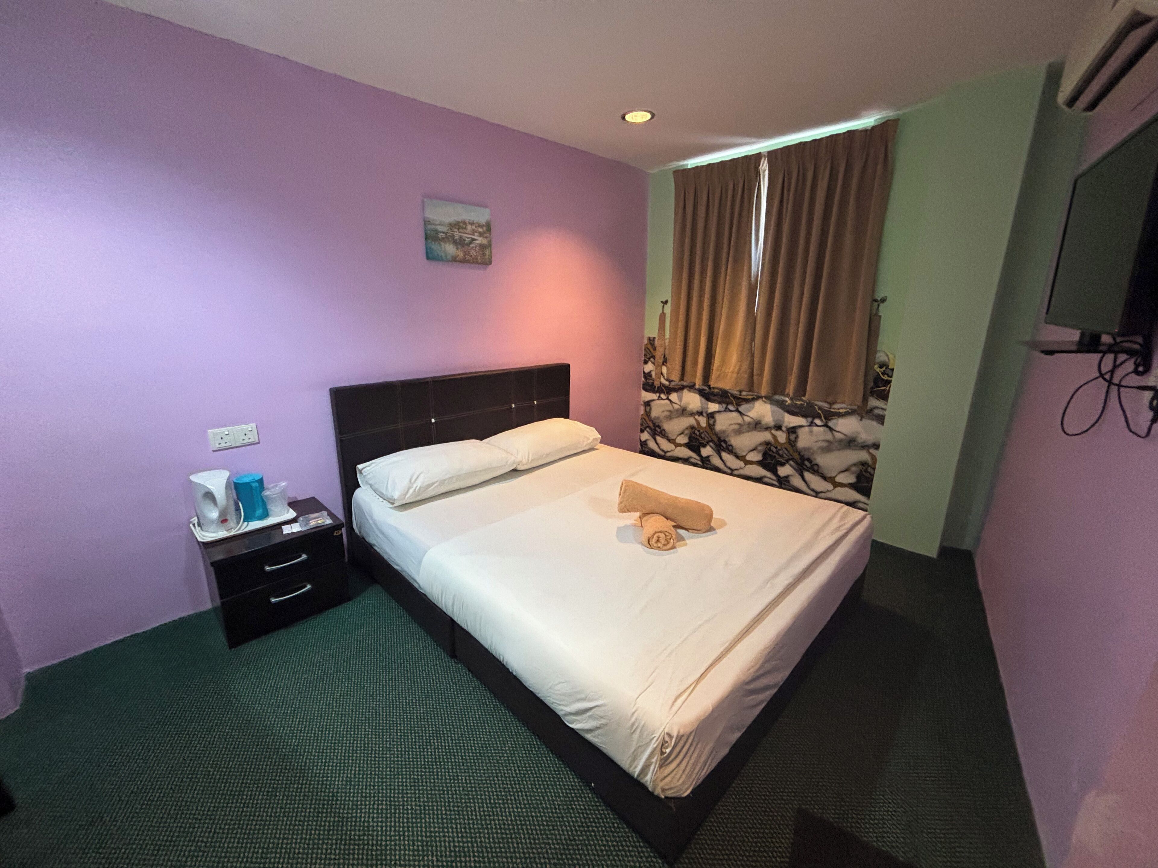 A hotel room with a bed, a nightstand, a small table with a lamp, a painting on the wall, and a window with curtains.