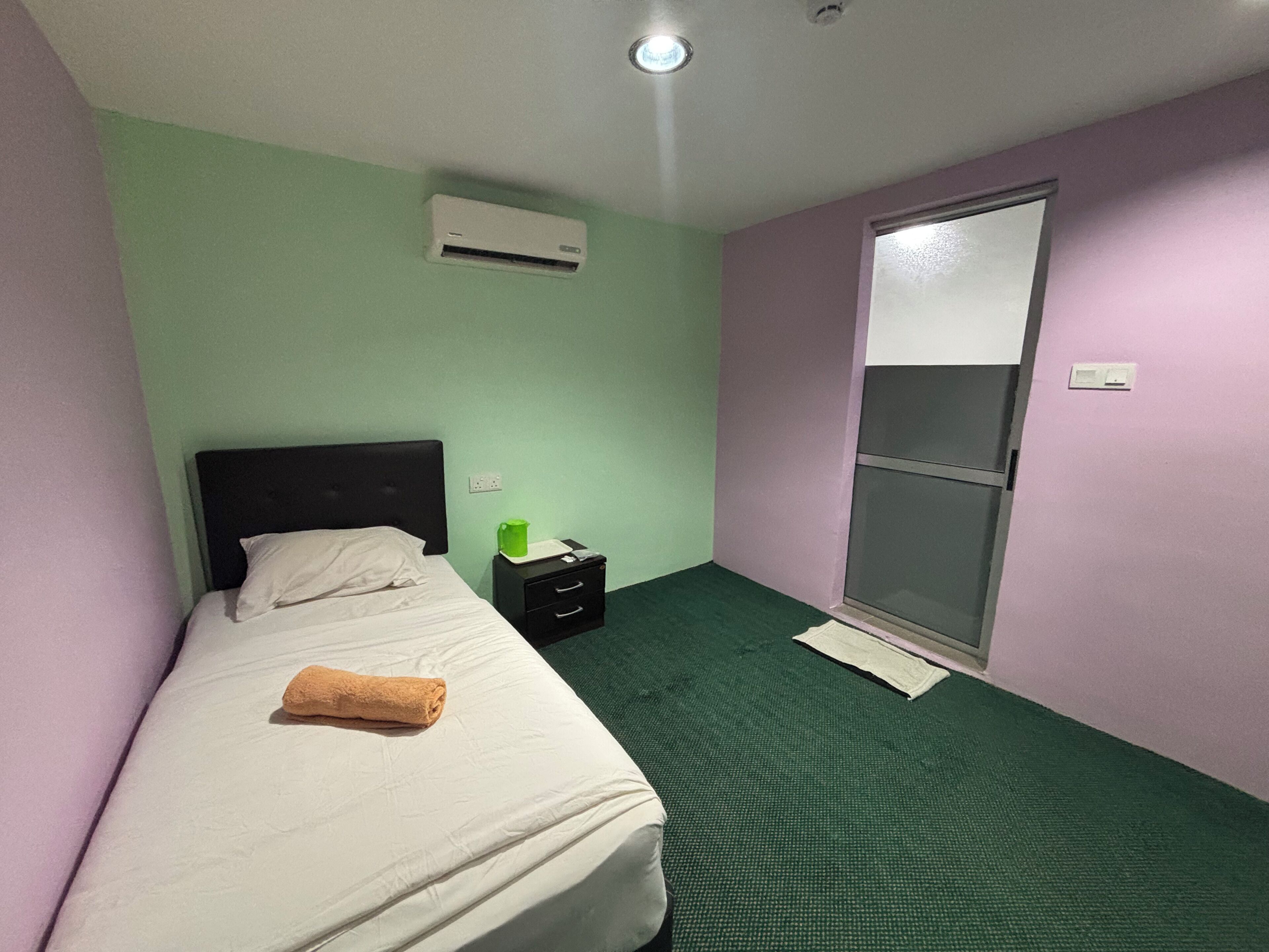 A single bed with a white mattress and a brown towel, a nightstand with a green cup, a black drawer, and a window with a blind.