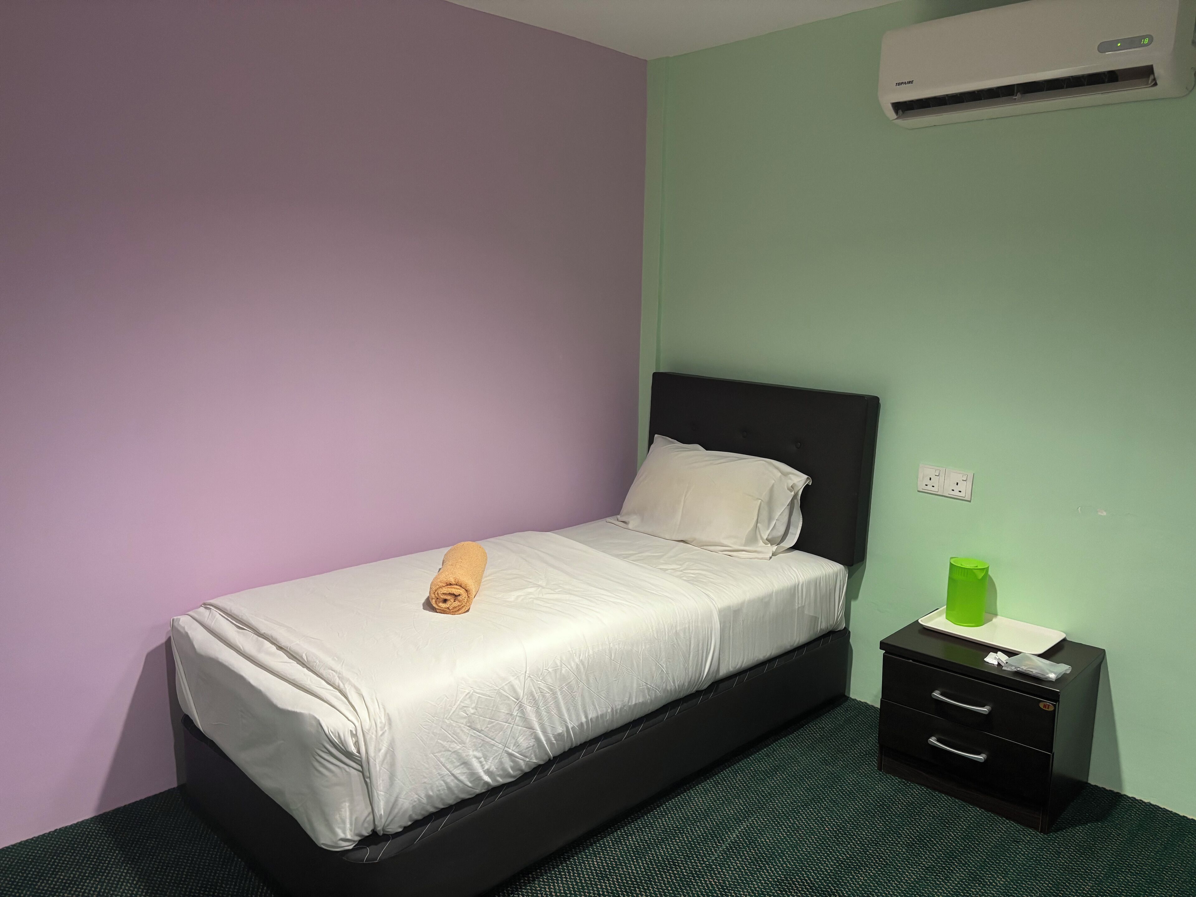 A single bed with a white mattress and a rolled towel, a black nightstand with a green bottle, and an air conditioning unit on the wall.