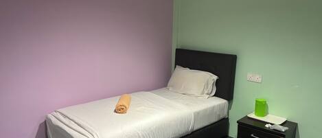 Un lit simple avec un matelas blanc et une serviette enroulée, une table de chevet noire avec une bouteille verte, et un appareil de climatisation fixé au mur.