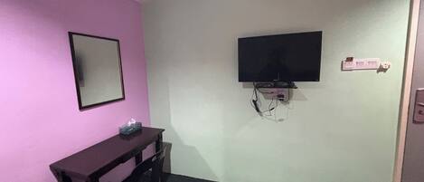 Una habitación con una cama, un escritorio, una silla, un televisor montado en la pared y un espejo en la pared.