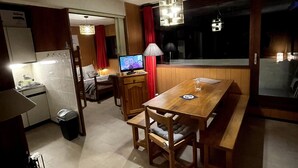 Un espacio habitable compacto con kitchenette, una zona de comedor con mesa y bancos de madera, televisor y un ventanal grande.