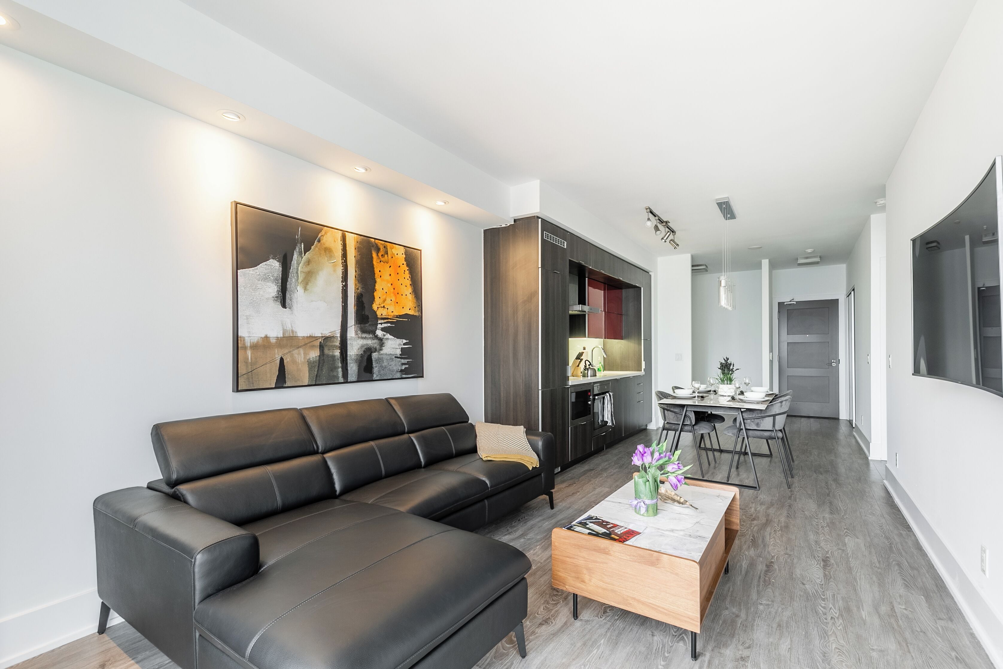 A modern living room with a black leather sofa, a wooden coffee table, and a dining area with a table set for four.