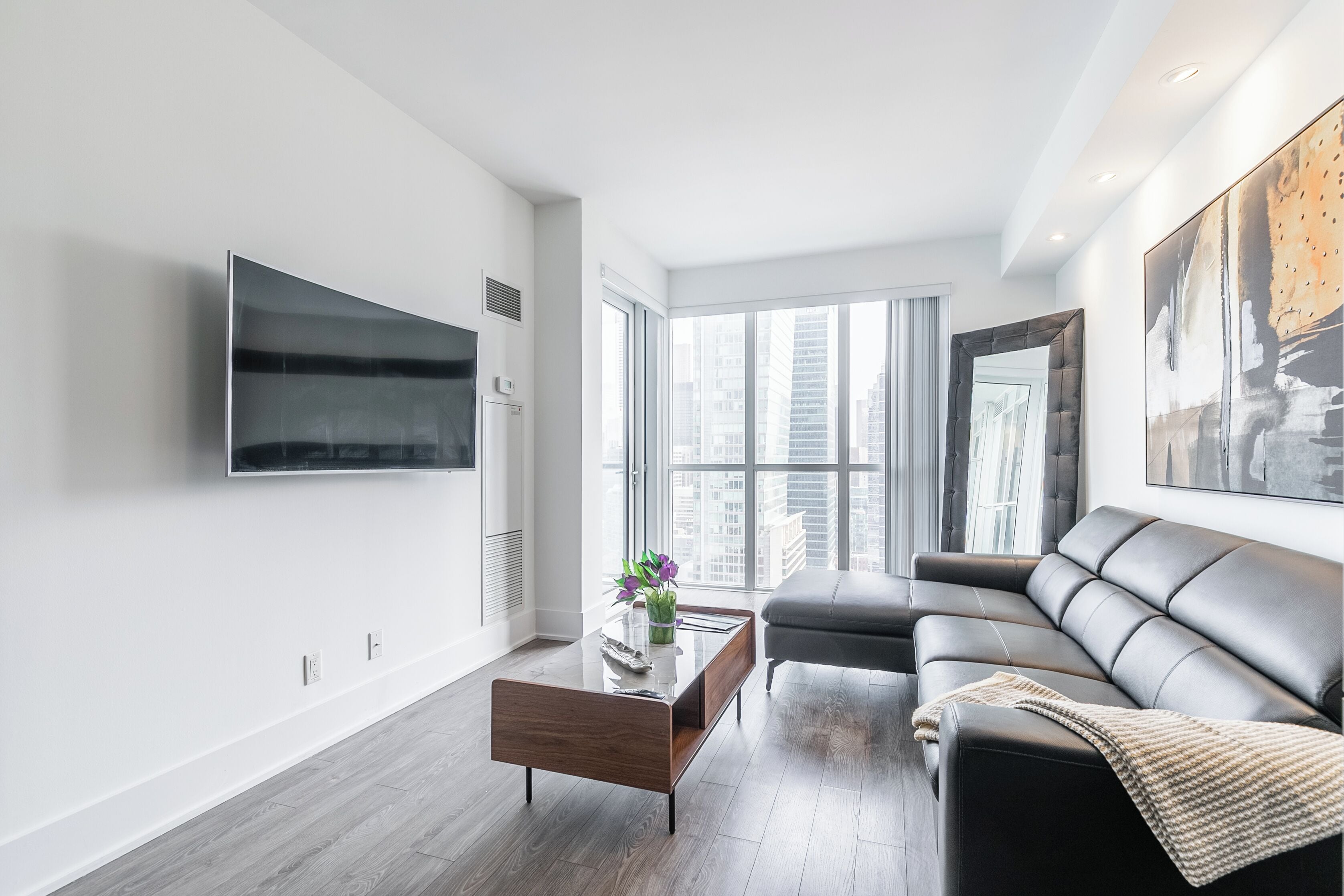 A modern living room with a flat-screen TV mounted on the wall, a leather sofa, a wooden coffee table, and a view of the city through large windows.