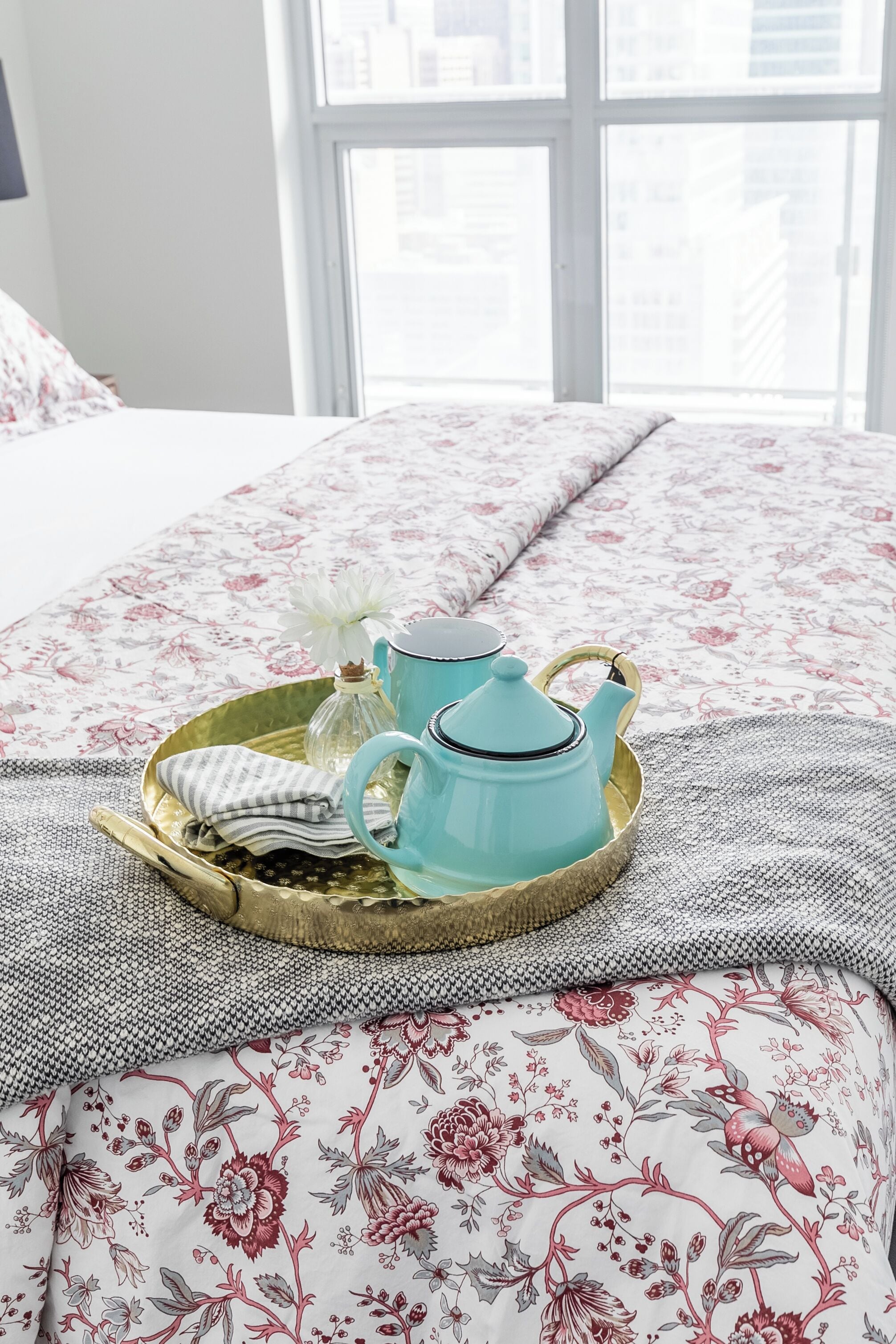Un letto con una coperta a motivi floreali, un vassoio con teiera e tazze, e una finestra con vista sugli edifici.