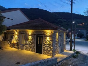 A stone building with a tiled roof, illuminated by exterior lights, situated on a paved street with a hilly landscape in the background. - VIDO STONE HOUSE (Kalavrita)