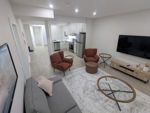 A modern living room with a grey sofa, two brown armchairs, a coffee table, and a TV on a wooden stand. - {Greystone Retreat} Walk to River, Downtown, Rec Center (Cochrane)