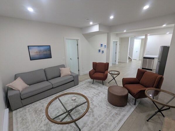 A modern living room with a grey sofa, two brown armchairs, a glass coffee table, and a patterned rug. - {Greystone Retreat} Walk to River, Downtown, Rec Center (Cochrane)