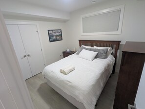 A neatly arranged bedroom with a bed, a bedside table, a nightstand, a wardrobe, a door, and a window with blinds. - {Greystone Retreat} Walk to River, Downtown, Rec Center (Cochrane)
