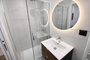 Ein modernes Badezimmer mit einer einachig verglasten Dusche, einem weißen Waschbecken und einem runden Spiegel mit integrierter Beleuchtung.