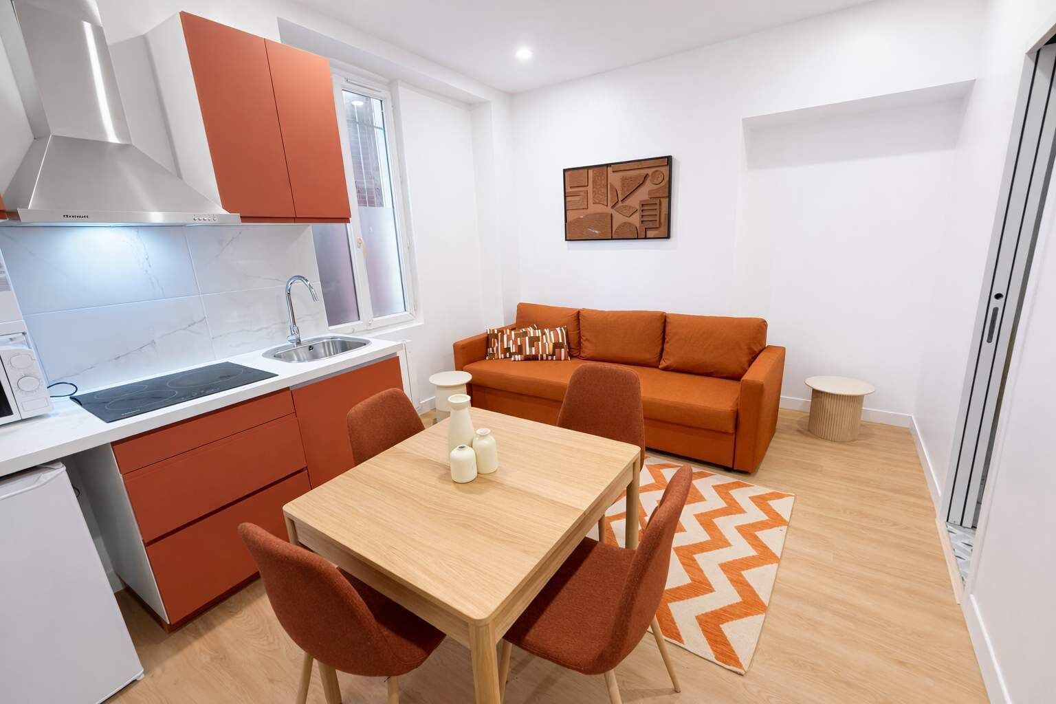Een compacte leefruimte met een kitchenette, een eettafel met stoelen, een bank en een kleine ronde tafel.