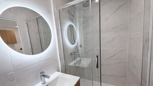 A modern bathroom with a glass-enclosed shower, a round mirror, and a white sink. - Appartement Cosy a Aubervilliers - 4P (Aubervilliers)