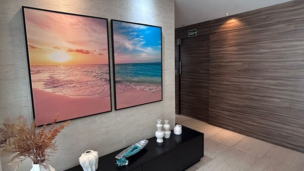 A modern lobby with a large wall-mounted TV displaying a sunset scene, a black console with decorative items, and a wooden panel wall. - AP ENCANTADOR 1 QUARTO PX. DO CAU HANSEN (Joinville)
