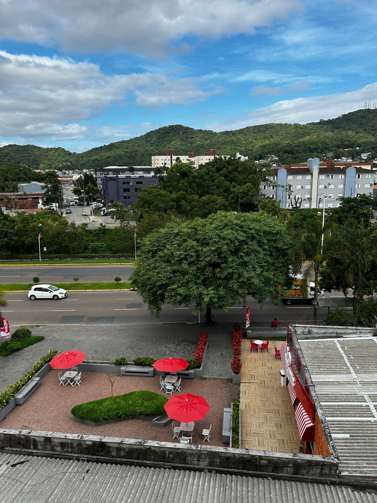 A rooftop terrace with red umbrellas and seating, overlooking a parking area and a road with vehicles.