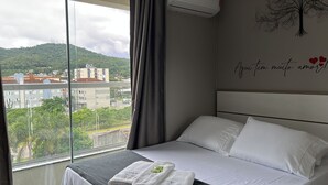 A hotel room with a bed, a window with a view of buildings and trees, a wall with a tree and hearts, and a curtain. - AP ENCANTADOR 1 QUARTO PX. DO CAU HANSEN (Joinville)