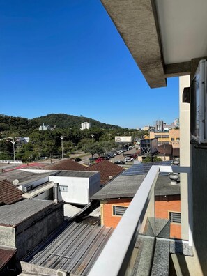 Kilátás egy erkélyről, ahonnan egy városképre nyílik rálátás: épületek, egy parkoló és a távolban egy domb látható.