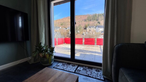 A room with a large sliding glass door leading to an outdoor area with a red fence. There is a flat-screen TV mounted on the wall, a wooden coffee table, and two potted plants. - Friedas Ferienhaus in Olsberg-bruchhausen (Olsberg)