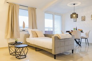 A modern living room with a sofa, a dining table, and chairs. There is a window with curtains, a pendant light, and a small table with a plant. - Neo Apartments - Modern 2-room apartment - free parking (Rastatt)
