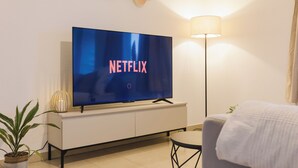 Moderni olohuone, jossa on seinälle asennettu taulutelevisio, jossa näkyy Netflixin logo, valkoinen kaappi, lattiavalaisin ja ruukussa oleva huonekasvi.