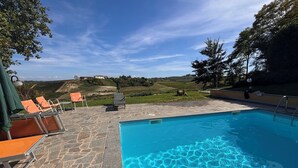 En swimmingpool med stentilfliser, orange liggestole og udsigt til et bakket landskab.